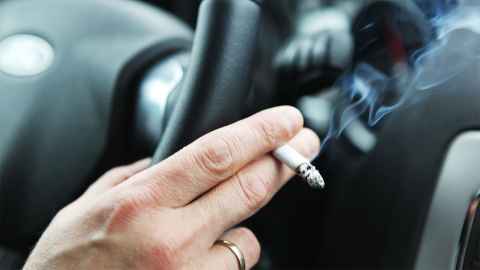 A hand of a car driver holding the steering wheel and a smoldering cigarette is shown: A submission opposing a ban on smoking in cars failed to disclose the link with funding from big tobacco. Photo: iStock 