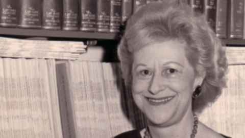 Black-and-white photo of Marylyn Mayo standing in front of shelves stacked with bound and unbound journals.