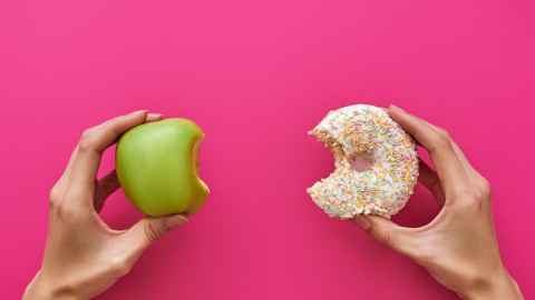 An apple with a bite out of it held in one hand, and a donut with a bite out of it in the other hand.