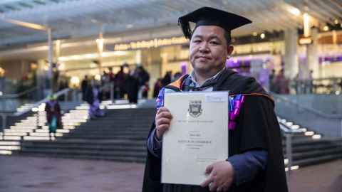 Bill Shen, wearing academic regalia and holding a degree certificate, standing outside the Aotea Centre.