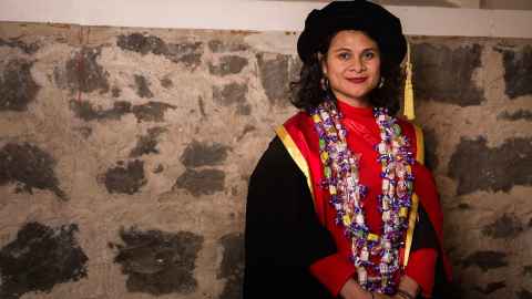 Dr Karamia Müller wearing academic regalia and standing next to a stone wall