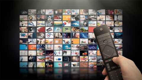 A large TV screen displays multiple video streaming site and a remote in the hand of an unseen user is in the foreground: Downstream traffic on the internet today is dominated by video-streaming. Photo: iStock