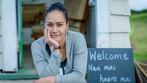 Pania Newton, sitting outside an open door with a sign reading "Welcome, Nau mai, haere mai"