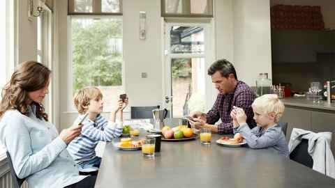 A family sit around a kitchen table, each totally absorbed in their mobile phones: A ‘healthy’ amount of social media use for adults isn't necessarily ‘healthy’ for their kids. Photo: iStock kupicoo