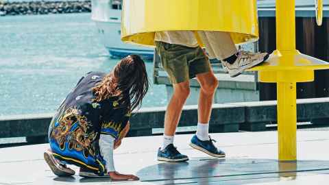 The feet and legs of two people emerging from a large yellow tube on a waterfront location, with a third person looking on.
