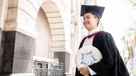 Eunice Ng standing outside Auckland Town Hall, wearing academic regalia and holding a frisbee