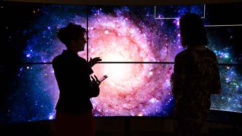Dr Héloïse Stevance (left) and Dr JJ Eldridge in the Centre for eResearch Visualisation Suite in front of representations of an image of a galaxy.