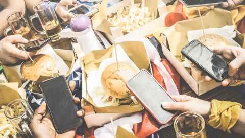 Five people using their phones at a table full of hamburgers and fries.