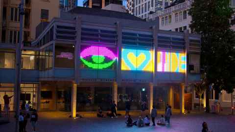 People sitting and standing in Freyberg Place watching the brightly coloured LED lights in the windows of the Ellen Melville Centre