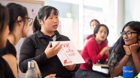 A student holding up a sheet of paper and speaking while other students look on.