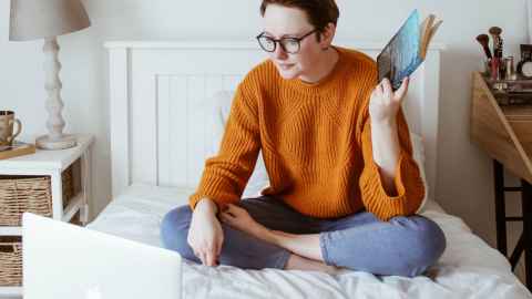 A person sitting cross-legged on a bed, looking at a laptop and holding a book in one hand.