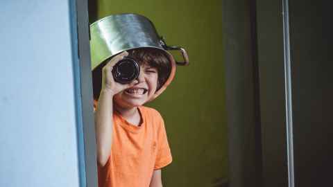 Child wearing a cooking pot on its head and looking through a bottle as a telescope