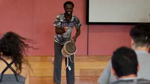 Dr Alfdaniels Mabingo standing and playing a drum held between his legs.