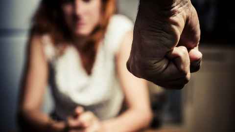 A woman sits at a table in the background with a clenched male fist in the foreground: The victim must be kept front and centre in work to tackle domestic violence. Photo: iStock