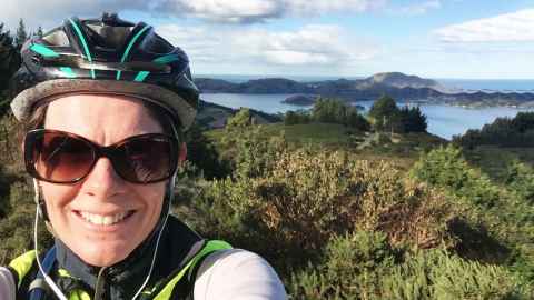 A selfie of a woman in a bike helmet with trees, green hills and water in the background.