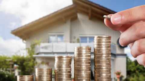 A hand adding to stacks of coins in front of a house.