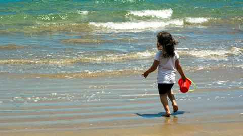 A little girl on a beach, at the water's edge, holding a bucket.