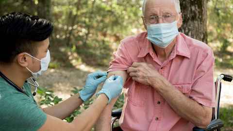 The image shows an elderly man receiving a vaccination from a young male health professional. Both are wearing face masks. Photo: iStock