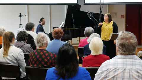 A man at a piano and a woman standing in front of a group of people.