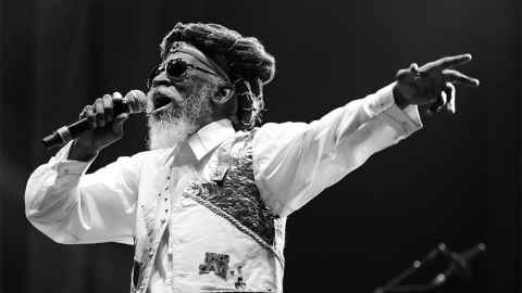 The image shows a recent image of Bunny Wailer singing on stage: Bunny walked this world as a proud Rastafarian and, despite some digressions into disco and dancehall, he remained identified with roots reggae and his Rastafarian beliefs. Photo: Peter Verwimp, CC0, via Wikimedia Commons