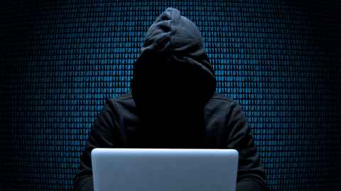 A person wearing a hoodie, in shadow, at a laptop with binary code on the wall behind them.