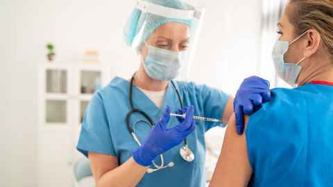 A woman in scrubs and PPE, giving another woman (in a surgical mask) an injection.