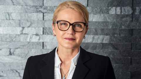 Dame Helen Winkelmann wears glasses and stands in front of a grey brick wall.