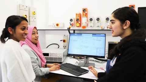 Three young women standing around a computer, talking.