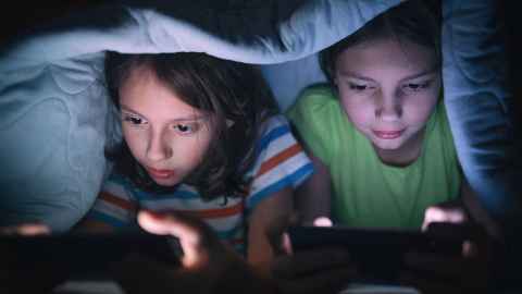 The image shows two young children secretly playing video games under the bed covers: The American Psychiatric Association 2013 defines Internet Gaming Disorder as “a persistent and recurrent use of the Internet to engage in games”. Photo: iStock