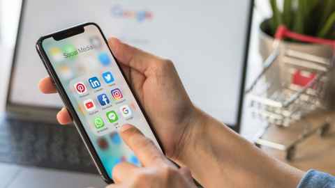 A person using a smartphone with a laptop open behind it with Google search on screen.