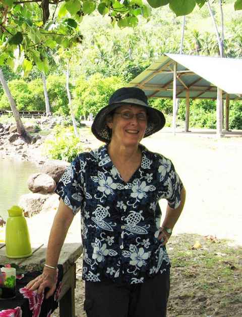 A woman in a hat and tropical-print shirt, with palm trees in the background.