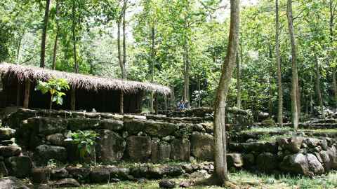 A rustic building with a thatched roof surrounded by stone walls and green trees.