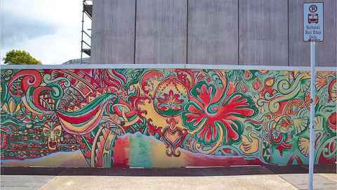 A red, green and yellow mural, with stylised flowers, leaves, faces and koru.