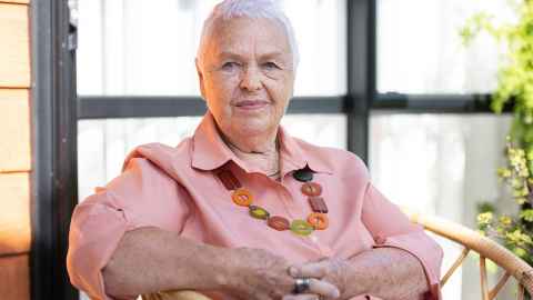  Judy Lessing visiting Auckland, sitting in a chair wearing pink shirt and a bright beads necklace. 