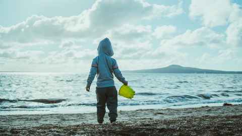 A child wearing a hoodie, looking out too Rangitoto from an Auckland beach.