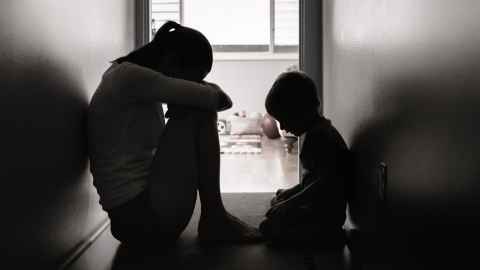 A woman and child sitting on the floor in shadow, leaning against the walls of a narrow hallway.