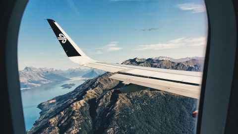 The view from an aeroplane window showing a wing with the Air NZ logo, mountains, lakes and blue sky.