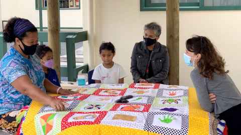 Women and children sitting around a table with a large piece of embroidered fabric on it.