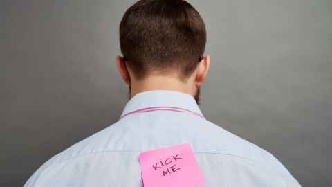 A man with a pink post-it note on his back that says 'kick me'.