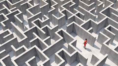 A grey concrete maze from above, with a woman in an orange dress attempting to find her way through it.