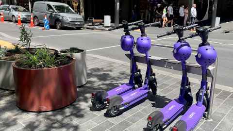 Four purple Beam scooters lined up on the footpath of a busy city street.