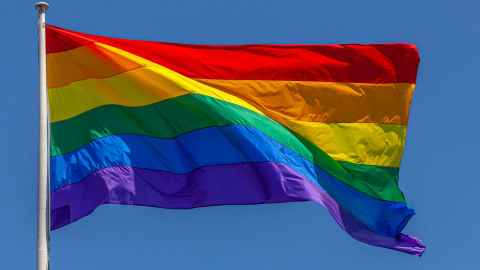 A rainbow flag flying in the wind, with blue sky in the background.