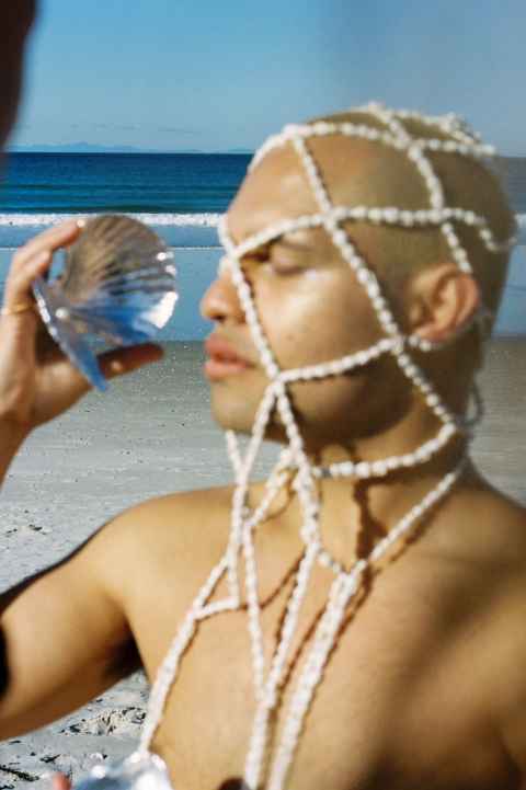 A man on a beach with a silver shell and beads over his face.
