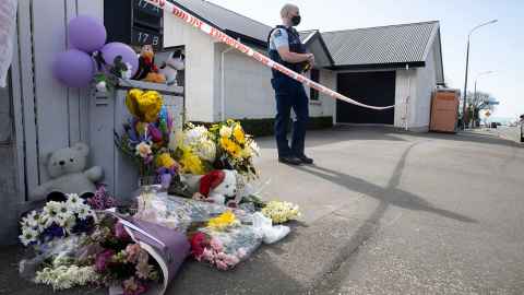 A house with police tape and a police guard at the front and flowers and teddy bears left by the letterbox.