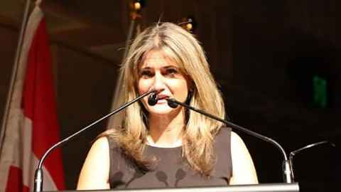 A woman at a microphone, with flags in the background.