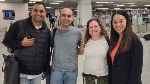 Two men on the left and two women on the right standing together at an airport smiling.