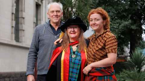 A bespectacled woman, a man, and a younger woman smiling at the camera. The bespectacled woman is wearing her doctoral regalia. 
