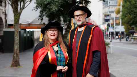 A bespectacled woman and a man smiling at the camera in their doctoral regalia. 