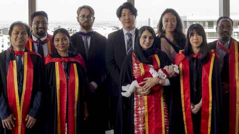 Group of students in graduation regalia in front of window and cityscape