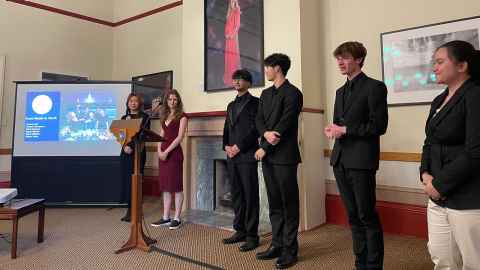 Six students in business attire standing to the side of a presentation screen